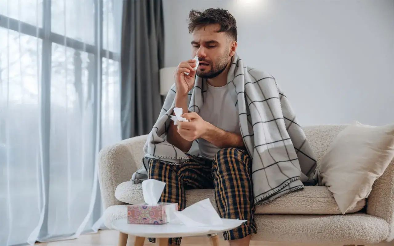 Mann mit Taschentuch und Nasenspray, eingewickelt in eine Decke. Er nimmt sich freizeitausgleich krank und erholt sich zu Hause.