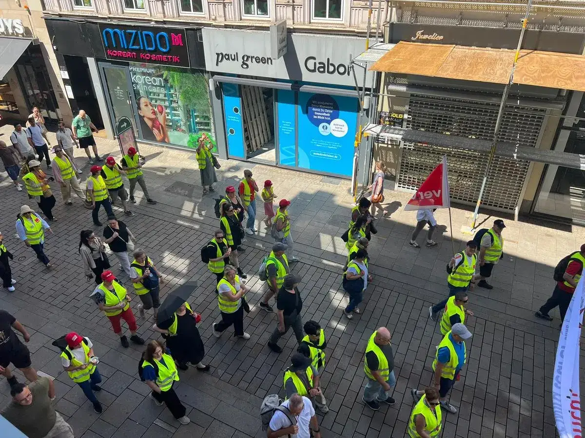 Menschen in Warnwesten marschieren auf einer Straße, einige tragen Fahnen. Ein Streik im Handel ist deutlich erkennbar.